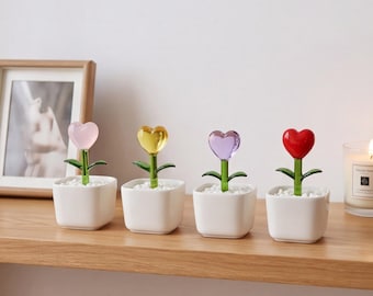 May include: Four small white square ceramic pots each containing a single glass flower with a heart-shaped bloom. The flowers have green stems and leaves, with blooms in translucent pink, yellow, purple, and solid red, set in white pebbles.