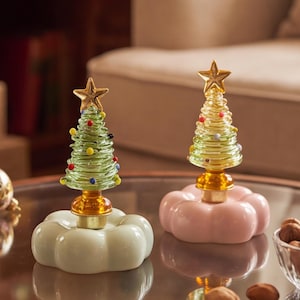 May include: Two glass Christmas tree figurines, one light green and one yellow, each topped with a gold star and decorated with small colorful glass spheres. The trees are mounted on rounded, scalloped bases in pale green and soft pink colors.