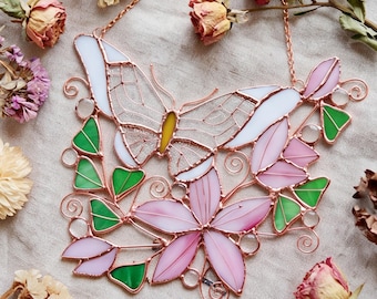 Puede incluir: Un atrapasoles de vidrieras que presenta una mariposa con alas blancas y cuerpo amarillo, rodeada de pétalos de flores rosas y hojas verdes. La pieza está construida con líneas de soldadura de metal cobrizo e incluye espirales de alambre decorativas.