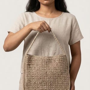 May include: A beige, rectangular handbag with a woven texture. The bag has a matching woven handle and is held by a person. The bag is made of a natural fiber material.
