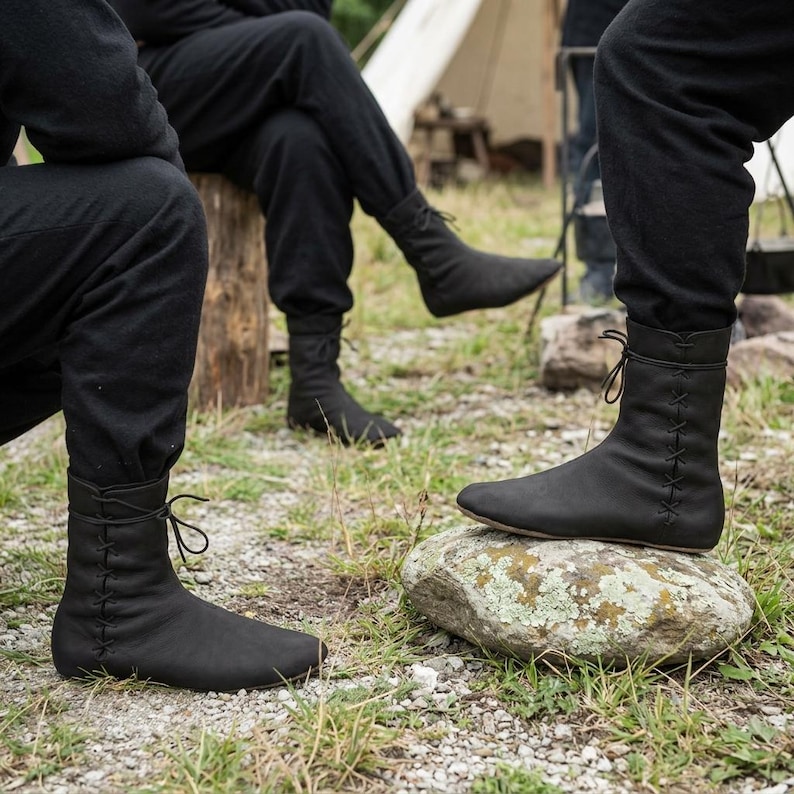 May include: A pair of black leather boots featuring a mid-calf height, a rounded toe, and a vertical lace-up closure along the outer side. The boots are constructed with a smooth matte finish and thin black laces tied at the top of the shaft.