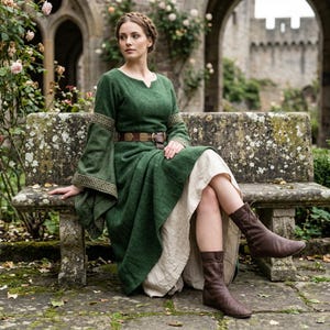 May include: A long-sleeved forest green dress with a notched neckline and wide sleeves featuring intricate gold-toned woven trim. The dress is worn over a cream-coloured underskirt and is cinched at the waist with a brown leather belt with a metal buckle.