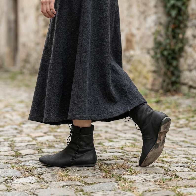May include: A dark grey ankle-length skirt with a textured surface paired with black leather ankle boots. The boots feature a rounded toe, a flat sole, and a vertical lace-up detail along the side, extending to the mid-calf with a slim fit.
