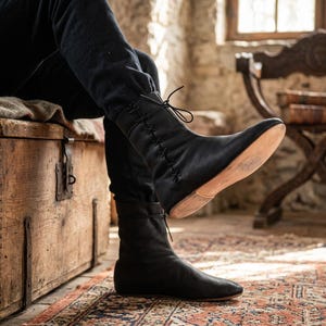 May include: A pair of black leather mid-calf boots featuring a lace-up front closure with thin black laces that crisscross vertically along the shaft. The boots have a rounded toe, a smooth matte finish, and a light tan flat sole visible on the bottom.