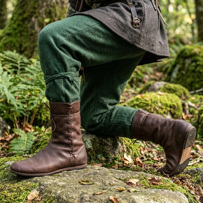 May include: A pair of dark brown leather boots featuring a side-lacing design with thin leather cords, paired with dark green trousers and a dark grey tunic with a leather belt, worn by an individual kneeling in a natural outdoor setting.