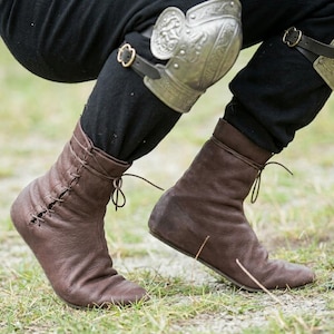May include: A pair of dark brown leather ankle boots with side lacing and thin leather ties, worn with black trousers and a silver metal knee guard featuring an embossed design and a black leather strap with a gold-toned metal buckle.