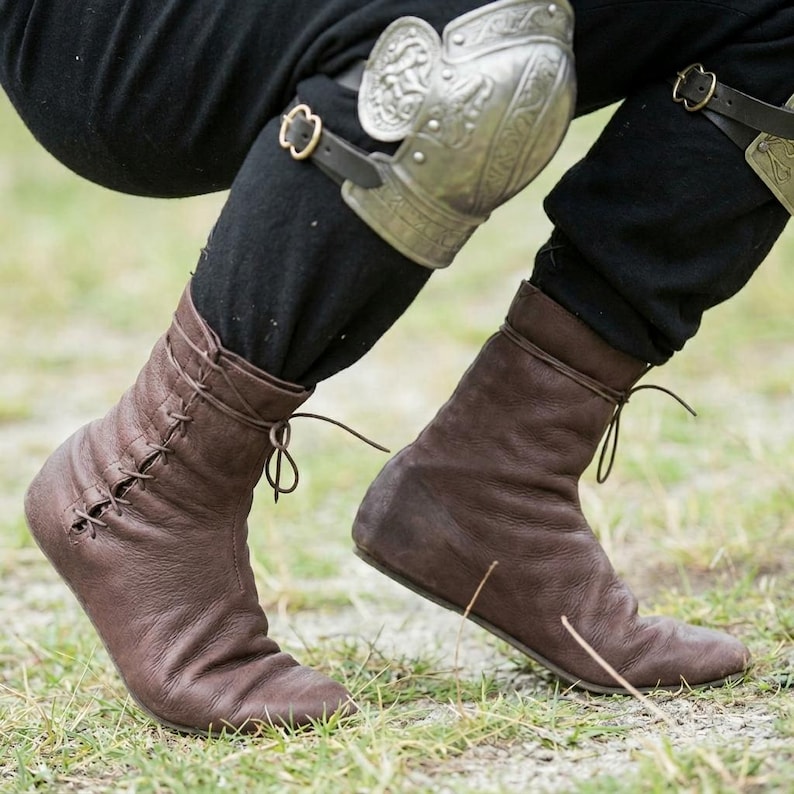 May include: A pair of dark brown leather ankle boots with side lacing and thin leather ties, worn with black trousers and a silver metal knee guard featuring an embossed design and a black leather strap with a gold-toned metal buckle.