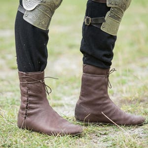 May include: A pair of brown leather ankle boots featuring side lacing with thin leather cords. The boots are worn over black trousers, with metallic, engraved knee-length armour plates secured by dark leather straps with buckles visible above.