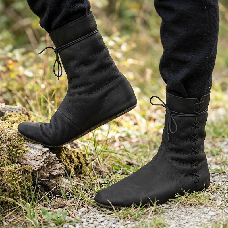 May include: A pair of black leather mid-calf boots featuring a rounded toe, a flat sole, and a side lacing system with thin black cords that tie at the top. The boots have a smooth matte finish and a flexible construction that contours to the foot.