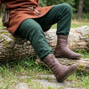 May include: A pair of brown leather mid-calf boots featuring a lace-up front closure with thin leather ties. The boots are worn with dark green wool trousers and a rust-coloured tunic, with a small brown leather pouch attached to a belt.