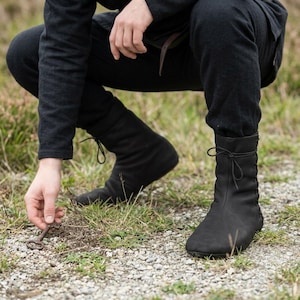 May include: A pair of black ankle boots featuring a soft, matte finish, rounded toes, and a side-lacing system secured with thin black laces tied at the top. The boots have a slim profile and a flexible construction, reaching just above the ankle.