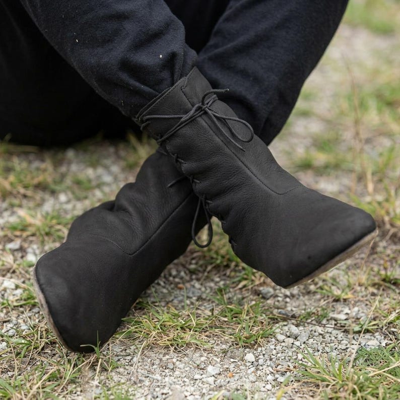 May include: A pair of black ankle boots featuring a soft, matte leather-like texture, a rounded toe, and a lace-up closure system that runs along the front of the shaft, tied in a bow at the top with thin, matching black laces.