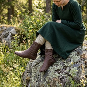 May include: A dark green long-sleeved dress with a notched neckline paired with beige knit leggings and brown leather ankle boots featuring side lacing details. The boots have a rounded toe and a flat sole, extending to the lower calf area.