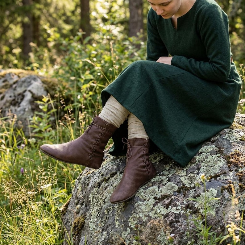 May include: A dark green long-sleeved dress with a notched neckline paired with beige knit leggings and brown leather ankle boots featuring side lacing details. The boots have a rounded toe and a flat sole, extending to the lower calf area.