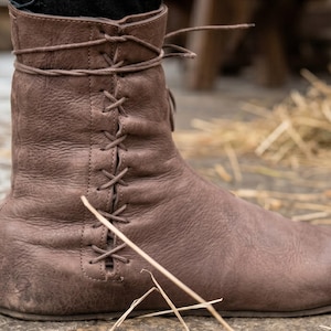 May include: A brown ankle boot with a textured surface and a side-lacing design featuring thin leather laces that crisscross through vertical eyelets along the outer side of the shaft, extending to a wrap-around tie at the top of the boot collar.