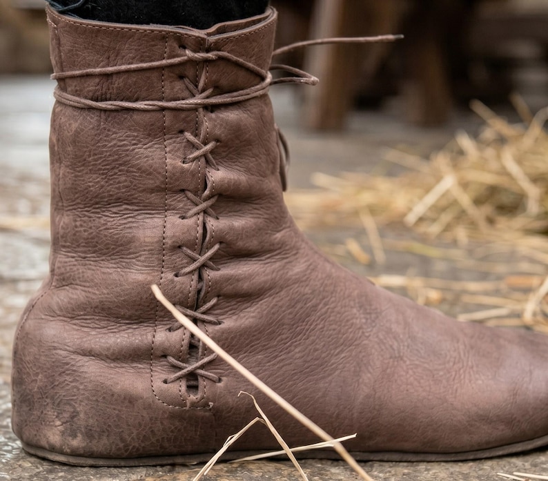 May include: A brown ankle boot with a textured surface and a side-lacing design featuring thin leather laces that crisscross through vertical eyelets along the outer side of the shaft, extending to a wrap-around tie at the top of the boot collar.