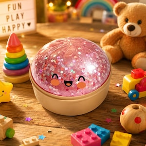 Könnte beinhalten: Ein kuppelförmiges sensorisches Spielzeug mit einer rosa, glitzergefüllten Oberseite und einer beigen Basis. Die Oberseite zeigt ein aufgedrucktes Cartoon-Gesicht mit schwarzen Augen, einem lächelnden Mund und orangefarbenen Wangen.