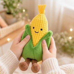Puede incluir: Un juguete de peluche con forma de mazorca de maíz sonriente, con granos amarillos y hojas verdes. El juguete tiene pies marrones y un mechón amarillo. Se sostiene en las manos.