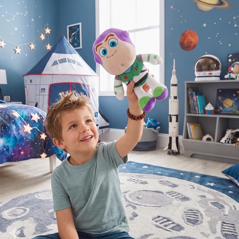 May include: A Buzz Lightyear soft toy from the Toy Story franchise. The toy is held up in the air by a person. The toy has a purple head, green and white body, and purple and green feet. The background includes a rocket ship, a moon rug, and a space-themed bedroom.