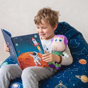 May include: A child reading a space-themed book with illustrations of rockets and astronauts. The child is holding a plush toy of a character in a green and purple space suit. The child is sitting on a blue beanbag chair with a constellation pattern.