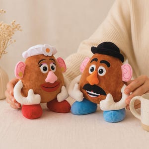 May include: Two plush potato-shaped toys, one with a white bonnet and flower, the other with a black hat and mustache. Both have large eyes, pink ears, and white gloves. The toys are brown with red, pink, and blue accents.