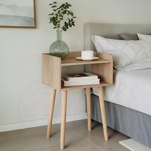 May include: A light wood bedside table with a two-tiered design. The top shelf holds a white teacup and saucer, while the lower shelf displays a stack of books. The stand has four angled legs and a vase with greenery. The nightstand is 61 cm tall.