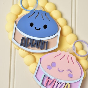 Puede incluir: Dos llaveros personalizados con forma de bollos al vapor en cestas de bambú. Un bollo azul lleva el nombre ADRIAN en letras azul oscuro. El bollo rosa lleva el nombre EMMA en letras multicolores. Ambos tienen anillas de plástico azul.