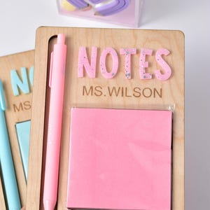Puede incluir: Organizador de escritorio de madera con la palabra "NOTES" en letras rosas y el nombre "MS. WILSON" debajo. Incluye un bolígrafo rosa y un bloc de notas adhesivas rosa. Un portalápices transparente con forma de corazón está en el fondo.