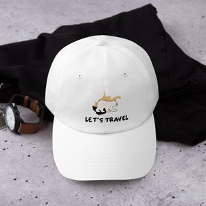 May include: White baseball cap featuring a bear and map design. The text "LET'S TRAVEL" is printed in black below the graphic. The cap has a curved peak and an adjustable strap.