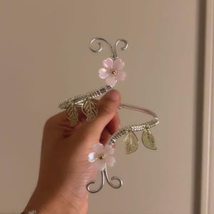 May include: A silver-toned wire arm cuff featuring two pale pink flower accents with gold centers. The design includes curled wire ends and four translucent green leaf charms attached to the spiraled metal band, creating a botanical aesthetic.