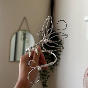 May include: A silver-toned wire ear cuff featuring an intricate wing-like design, accented with two small white pearls and two delicate metallic leaf charms. The structure is crafted from coiled wire, forming a curved shape that wraps around the ear.