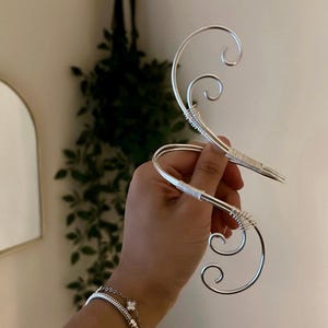 May include: A pair of silver-colored ear cuffs featuring a curved, swirling design with delicate wire-wrapped detailing at the center points. The metal structure forms an elongated, open shape that follows the contour of the ear.