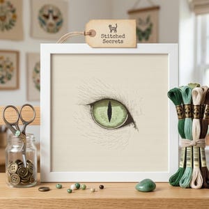 May include: A framed cross-stitch of a cat's eye, featuring a detailed green iris and white fur. The frame is white, and the artwork is displayed on a wooden surface with embroidery floss, scissors, and buttons. A tag reads "Stitched Secrets."