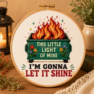 Könnte beinhalten: Ein runder Stickrahmen aus Holz mit einem Kreuzstichmotiv eines brennenden grünen Müllcontainers. Auf dem Container steht der Text THIS LITTLE LIGHT OF MINE und der untere Text lautet I'M GONNA LET IT SHINE in schwarzen und roten Buchstaben.