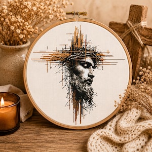 May include: A circular wooden embroidery hoop containing a cross-stitch pattern of a bearded man's face in profile, rendered in black and metallic gold thread. The design features abstract, linear geometric elements extending outward from the head.