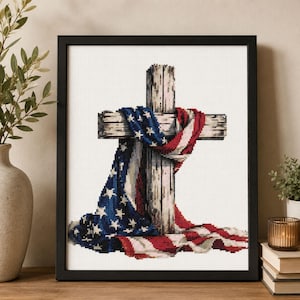 May include: A framed cross-stitch artwork featuring a weathered wooden cross draped with an American flag. The flag displays a blue field with white stars on the left and red and white stripes cascading down, all set against a white grid background.