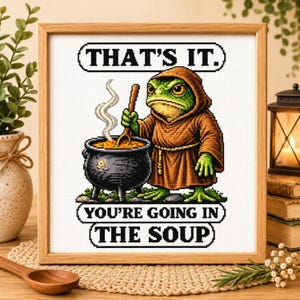 Könnte beinhalten: Ein gerahmtes Kreuzstichbild mit einem grünen Frosch in einer braunen Kapuzenrobe, der neben einem schwarzen Kessel mit einem Holzlöffel steht. Der Text lautet THAT'S IT. YOU'RE GOING IN THE SOUP in schwarzen Großbuchstaben auf weißem Stoff.