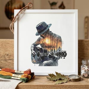May include: A square white frame containing a cross-stitch embroidery of a person wearing a hat and playing an acoustic guitar. The figure is filled with a double-exposure style landscape scene featuring silhouetted trees and a bright orange sunset.