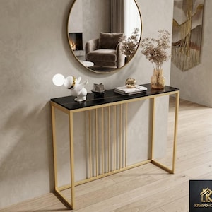 May include: A modern console table with a black marble top and gold metal frame. The table features vertical gold bars on the front and is decorated with a white sculpture, books, and a vase with dried flowers. A round mirror hangs above.