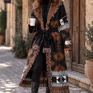 Peut inclure: Un long manteau ouvert orné d'un motif géométrique noir, marron et blanc. Le vêtement présente un large col châle, des manches longues et une ceinture en tissu noir à la taille. Les bords sont finis avec une épaisse bordure à franges.