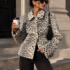 Pode incluir: Um casaco de manga comprida com base branca e um denso padrão de leopardo preto. O casaco tem um colarinho estruturado, fecho frontal com botões e uma cintura ajustada. É combinado com calças pretas e uma mala de ombro preta.
