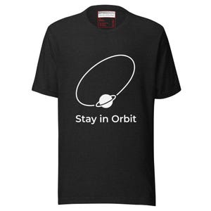 Puede incluir: Camiseta gris oscuro con un gráfico blanco de un planeta con un anillo y el texto "Stay in Orbit". La camiseta tiene cuello redondo y mangas cortas. La etiqueta de la marca es visible en la parte superior.
