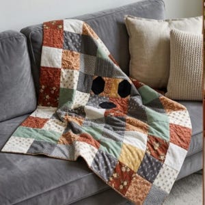 Może przedstawiać: Narzuta patchwork składająca się z siatki kwadratowych bloków materiału w odcieniach rdzawego pomarańczu, szałwiowej zieleni, musztardowej żółci, antracytowej szarości i kremu. Bloki prezentują różne wzory kwiatowe i geometryczne, połączone widocznymi szwami.