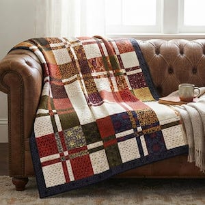 May include: A patchwork quilt featuring a grid of squares in shades of cream, deep red, olive green, and navy blue. The fabric squares display various intricate patterns, including floral and geometric motifs, finished with a dark navy blue border.