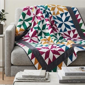 May include: A quilted throw blanket featuring a geometric star pattern in vibrant shades of magenta, teal, green, and mustard yellow on a white background, finished with a solid dark gray border and detailed stitching throughout the fabric.