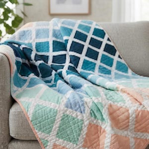 May include: A quilted throw blanket featuring a repeating diamond grid pattern in a gradient of colors, transitioning from deep navy and teal blue to light blue, mint green, and soft peach, finished with a solid peach-colored fabric edge binding.