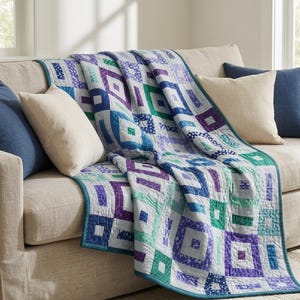 May include: A quilt featuring a geometric pattern of nested squares in shades of purple, teal, blue, and white. The fabric squares are arranged in a repeating block design with a solid teal border trim along the edges of the rectangular textile.