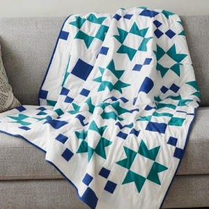 May include: A white quilt featuring a repeating geometric pattern of teal eight-pointed stars and solid blue squares. The textile has a blue fabric border along the edges and visible quilted stitching lines throughout the surface of the fabric.