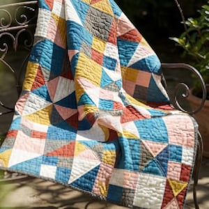 May include: A quilted blanket featuring a geometric patchwork pattern composed of triangular and square fabric segments in shades of mustard yellow, teal blue, terracotta orange, soft pink, white, and charcoal gray with visible stitching.