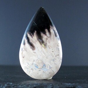 May include: A teardrop-shaped cabochon of agate with a black and white pattern. The agate has a white base with black and gray swirls and a black top.